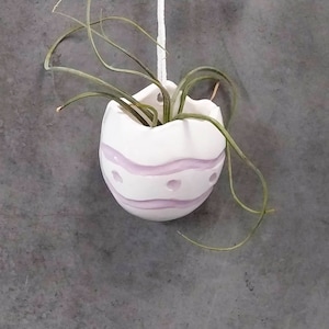 May include: White ceramic egg-shaped planter with pink stripes and holes, hanging from a white string, with a green air plant inside.