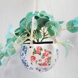 May include: A white ceramic hanging planter with a blue and white floral pattern. The planter is filled with green leafy plants and is hanging from a white string.
