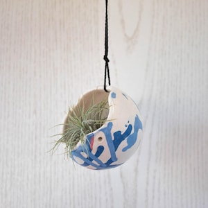 May include: A white ceramic hanging planter with blue and white abstract paint splatters. The planter has a hole in the side and is hanging from a black cord. A small air plant is growing inside the planter.