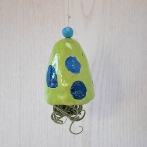 Puede incluir: Una campana de cerámica hecha a mano con un esmalte verde claro y grandes lunares azules. Una pequeña cuenta azul se encuentra en la parte superior de la campana, y zarcillos verdes y rizados cuelgan de la parte inferior. La campana está suspendida por un hilo fino.