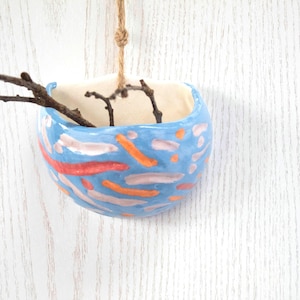 May include: A blue ceramic hanging planter with a colourful abstract design. The planter is decorated with pink, orange, and red lines and dots. The planter is hanging from a string and has a few small branches inside.