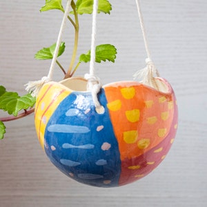 Op de afbeelding: Een kleurrijke keramische hangende plantenpot met een geometrisch ontwerp in blauw, geel, oranje en roze. De pot hangt aan witte koorden.