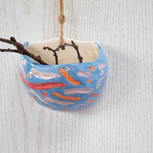 Op de afbeelding: Een blauwe keramische hangende plantenpot met een kleurrijk abstract ontwerp. De pot is versierd met oranje, roze en rode lijnen en stippen. De pot hangt aan een touwtje en heeft een paar kleine takjes erin.