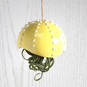 May include: A yellow ceramic planter shaped like a jellyfish with white polka dots and a green air plant hanging from it.