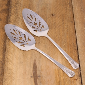 May include: Two silver cake servers with ornate cutouts in the shape of leaves and flowers. The servers have a polished finish and are laid out on a wooden surface.