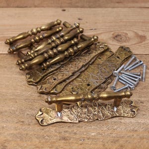 May include: A selection of antique brass door handles and pulls. The handles feature an intricate, decorative design with a warm, golden colour. Several matching backplates and screws are also visible, suggesting a complete set for restoration or new construction.