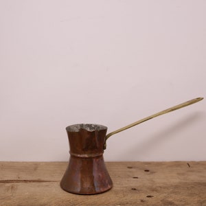 May include: A copper coffee pot with a brass handle. The pot has a rounded bottom and a flared top. It is sitting on a wooden surface.