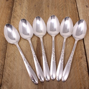 May include: Six silver spoons with an ornate floral design on the handle. The spoons are arranged in a semi-circle on a wooden surface.