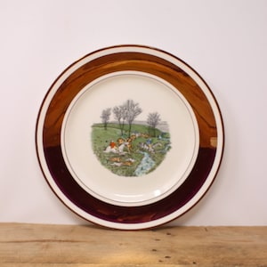 May include: A round decorative plate with a brown and burgundy rim. The center of the plate features a pastoral scene with figures, dogs, and trees. The plate is set on a wooden surface.