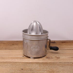 May include: A vintage, silver-toned citrus juicer with a ribbed reamer and a cylindrical base. It features a black handle for turning and a small emblem on the front. The juicer is made of metal and sits on a wooden surface.