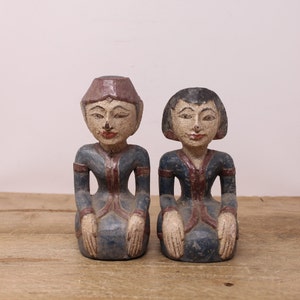 May include: Two wooden figurines of people in traditional clothing, kneeling with their hands clasped in front of them. The figurines are painted in shades of blue, red, and brown.