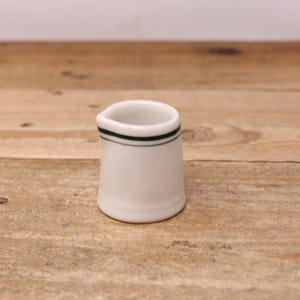 May include: A small white ceramic cream jug featuring a simple, thin dark green horizontal band encircling the upper portion of the body, just below the rim. The vessel has a smooth, glossy finish and a small integrated pouring spout.