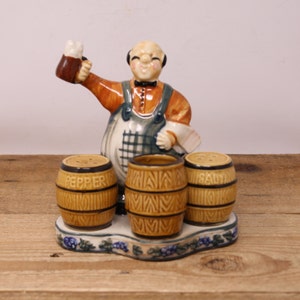 May include: Ceramic salt and pepper shaker set featuring a figurine of a man holding a beer mug. The shakers are shaped like barrels and have the words "Salt" and "Pepper" printed on them.