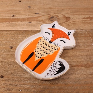 May include: A ceramic trinket dish shaped like a fox with a white base, orange accents, and black details. The fox features a patterned chest, closed eyes, and a gold-coloured rim. The design includes geometric shapes on the chest and tail.