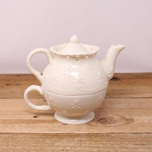 May include: Cream-colored ceramic teapot and teacup set. The teapot has a rounded body, a curved spout, and a handle. The lid is adorned with a small knob and decorative cutouts. The teacup is integrated into the base, featuring a handle and a footed base.