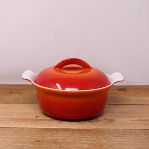 May include: A small, round, red ceramic casserole dish with a matching lid. The dish has white handles and a white rim. The lid has a curved handle. The dish is on a wooden surface.