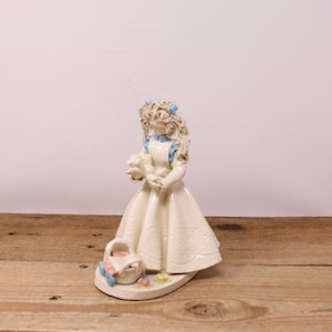 May include: A white ceramic figurine of a girl with blonde hair wearing a white dress and blue ribbon holding a white bird. The girl is standing on a white base with a basket and flowers.