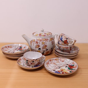 May include: A porcelain tea set featuring a teapot, three teacups, and four saucers. Each piece is decorated with hand-painted scenes of figures in colorful traditional clothing amidst cherry blossom trees with pink and yellow flowers.