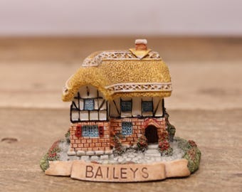 May include: A miniature thatched-roof cottage with brick walls and a sign that reads "BAILEYS". The cottage has a small door and windows, and is surrounded by greenery.