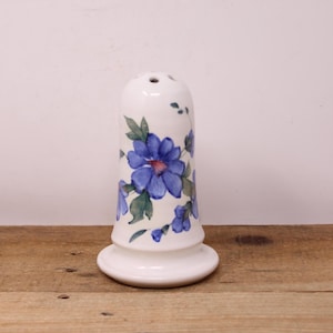 May include: A white ceramic salt shaker with a flared base and a rounded top with several holes. The shaker is decorated with blue floral designs and green leaves. The salt shaker is on a wooden surface.