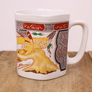 May include: White ceramic mug featuring a colourful design of a bird in flight, surrounded by stylised flowers and foliage. The design incorporates shades of red, yellow, green, and grey.