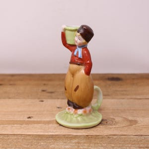 May include: A vintage ceramic figurine of a young person in traditional Dutch attire. The figure wears a red jacket, brown hat, and tan pants, carrying a green bucket. The figurine is mounted on a green base with floral accents.