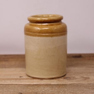 May include: A vintage stoneware jar with a cylindrical shape. The jar has a light beige body with a darker tan rim and a decorative band. The jar is on a wooden surface.
