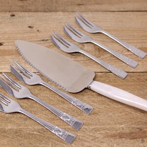 May include: A set of six silver cake forks and a silver cake server with a white handle. The forks and server have a decorative design.