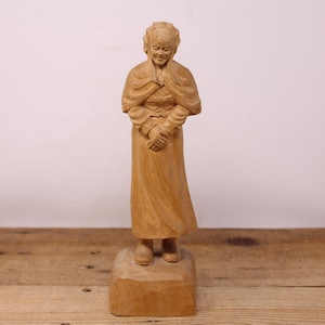 May include: A wooden carving of a woman in a long dress and a head covering. The carving is detailed and realistic, with a warm brown colour.