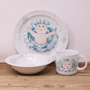 May include: A white ceramic baby dish set featuring a plate, bowl, and mug. Each piece is decorated with a baby design inside a blue cabbage. The plate and bowl have a dotted border.