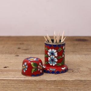 May include: A red enamel toothpick holder with a matching lid. The holder and lid are decorated with white and blue floral designs and green leaves. The holder contains several wooden toothpicks. The holder is on a wooden surface.
