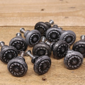 May include: A group of 14 silver-toned cabinet knobs with a decorative, circular design. The knobs have a distressed finish and are arranged in a pile on a wooden surface.