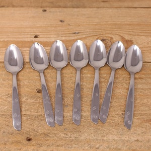 May include: Seven silver spoons with a star design on the handle. The spoons are arranged in a row on a wooden surface.