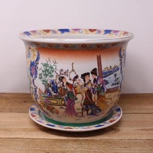 May include: A decorative ceramic planter with a matching saucer. The planter features a colourful illustration of figures in traditional attire, set against a landscape with mountains and trees. The rim and saucer have floral patterns.