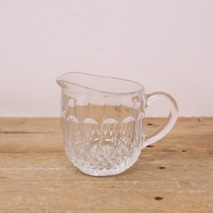 May include: A clear glass creamer with a diamond pattern. The creamer has a handle and a rounded base.