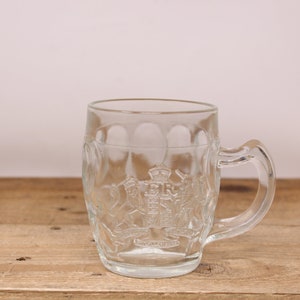 May include: A clear glass beer mug with a raised design of a crown and lions. The mug has a handle and is sitting on a wooden surface.