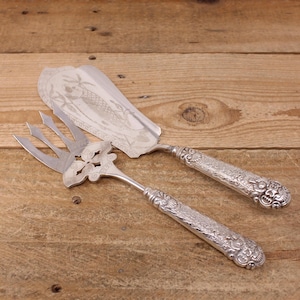 May include: Silver serving set featuring a cake server and a fork. The server has a fish design, and both pieces have ornate handles. The set is displayed on a wooden surface, showcasing the intricate details of the silverware.