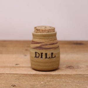 May include: A small, brown and tan ceramic jar with a cork lid. The jar is labeled "DILL" in black lettering.