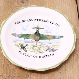 May include: A white ceramic plate with a green scalloped edge. The plate features a colorful illustration of three airplanes, including a green and brown Spitfire, and the text "THE 50th ANNIVERSARY OF THE BATTLE OF BRITAIN".