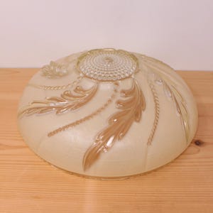May include: A dome-shaped glass light fixture shade with a frosted cream finish. The surface features embossed, raised leaf patterns in a light brown hue, accented by delicate beaded lines and a textured, circular centre medallion design.