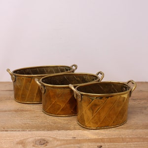 May include: Three oval-shaped brass planters with decorative handles. The planters are nested inside each other, with the largest planter at the bottom and the smallest planter at the top. The planters have a hammered finish and a warm gold colour.