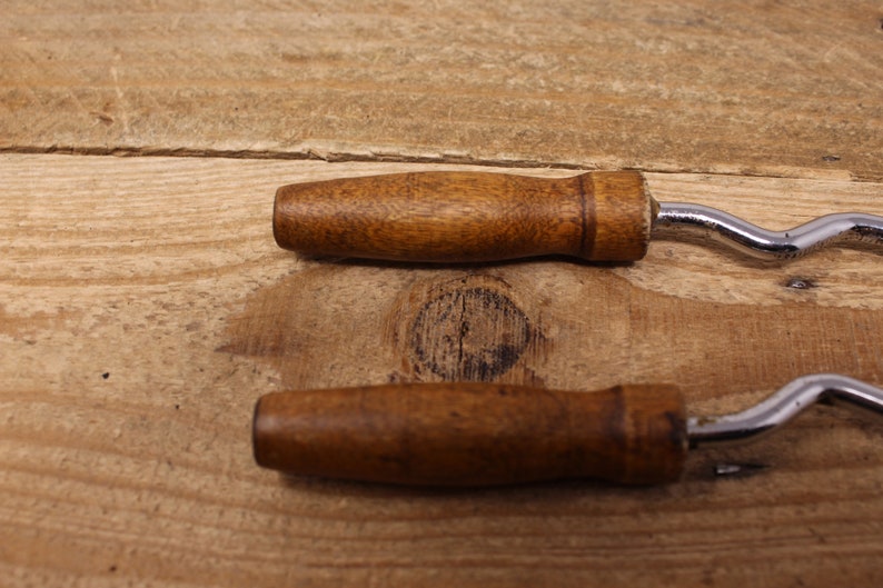 May include: Two vintage wooden handled bottle openers with a silver, wavy metal opener. The openers are on a wooden surface.