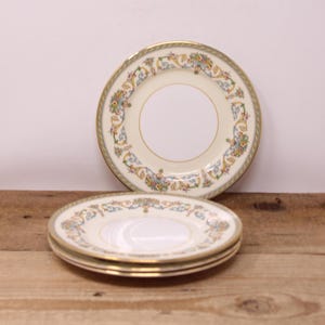 May include: A set of cream-coloured plates with a gold rim and floral design. The image shows a stack of three smaller plates and one larger plate. The plates have a cream-coloured centre and a decorative border with floral and scroll patterns.