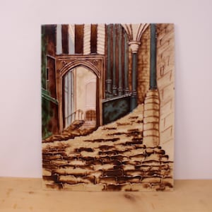 May include: A rectangular ceramic tile featuring a detailed relief illustration of a stone staircase leading towards a gothic archway. The scene is rendered in shades of brown, cream, and dark green, depicting architectural columns and textured masonry.