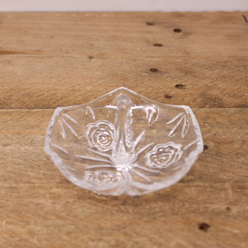 May include: A clear glass ring holder with a floral design and a handle. The holder is shaped like a leaf and has a delicate pattern of roses and leaves.