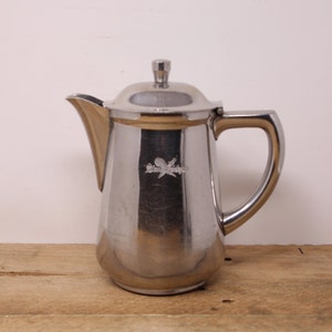 May include: A polished stainless steel coffee pot with a lid and handle. The pot features a spout and an emblem. It is displayed on a wooden surface.