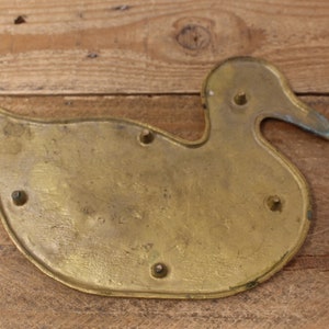 Pode incluir: Uma decora&ccedil;&atilde;o de parede em forma de pato de lat&atilde;o com um acabamento liso e brilhante. O pato tem um corpo arredondado e um pesco&ccedil;o longo e curvo. Existem cinco pequenos orif&iacute;cios na parte de tr&aacute;s do pato, provavelmente para montagem.