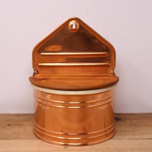 May include: A copper wall-mounted salt or sugar dispenser with a hinged lid and a white porcelain liner. The dispenser has a rounded shape and a decorative design.