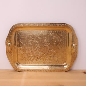 May include: A rectangular brass tray with rounded corners and two side handles. The surface features an intricate, engraved floral pattern covering the centre, while a decorative leaf motif borders the entire outer edge of the tray.