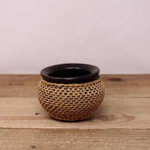 May include: A small, round, woven basket with a dark brown interior and rim. The basket's exterior is a light tan colour, and it sits on a wooden surface. The basket has a textured, open weave design.